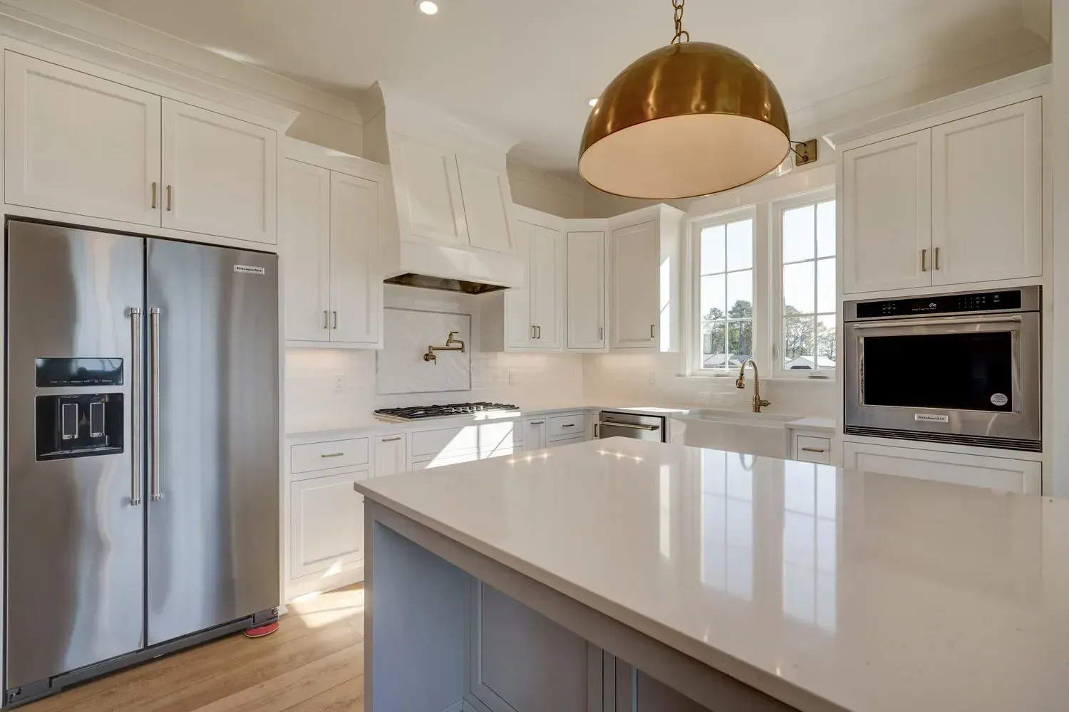 A bright kitchen with white cabinets, a large stainless steel refrigerator, white countertops, and a brass dome pendant.