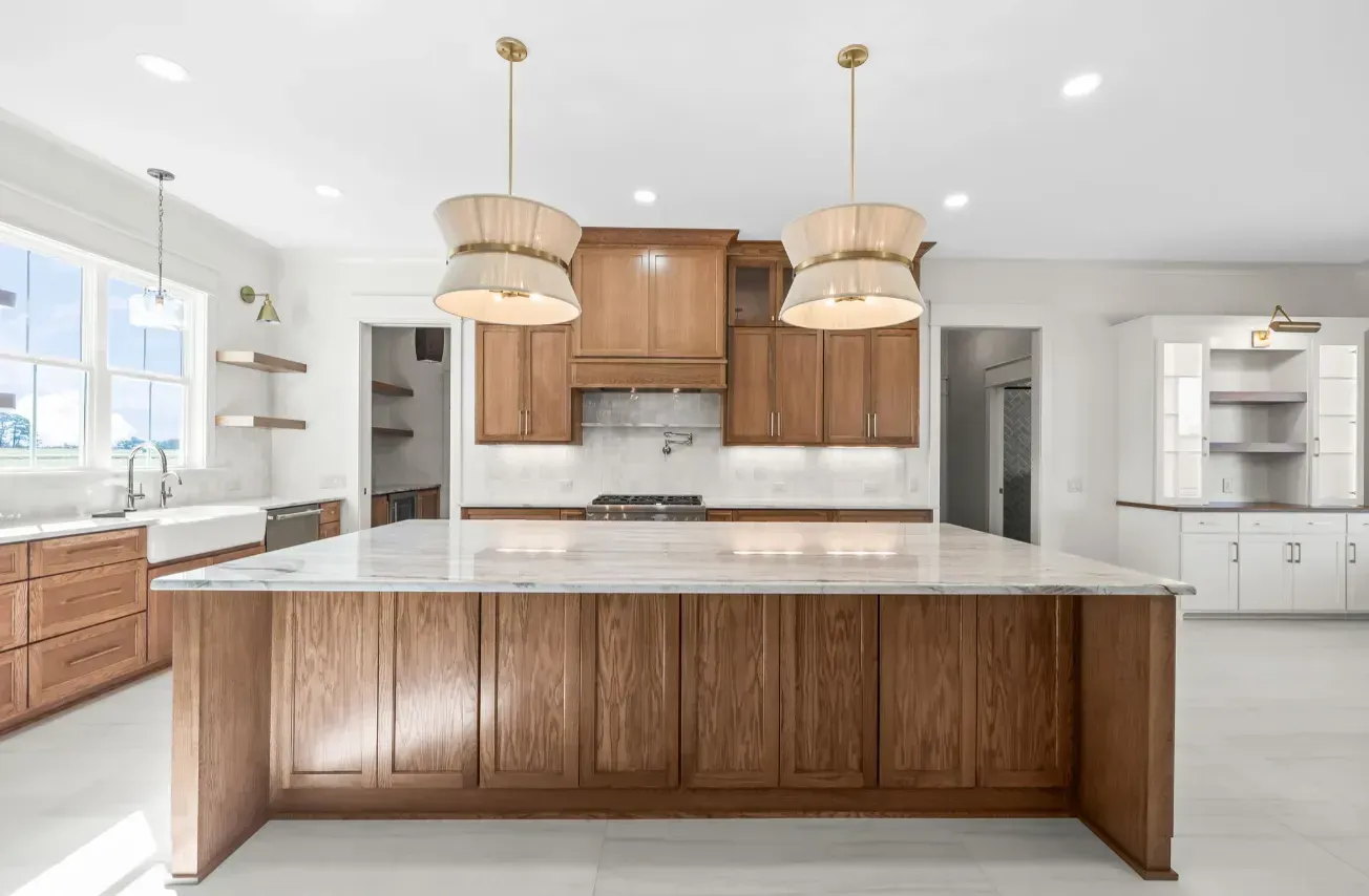 A large kitchen island with a white countertop sits before wood cabinetry, a stove, and two hanging pendant lights.