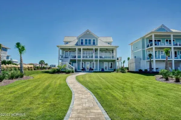 A wide view of a light blue beach house with a stone pathway leading through a green lawn under a clear blue sky.