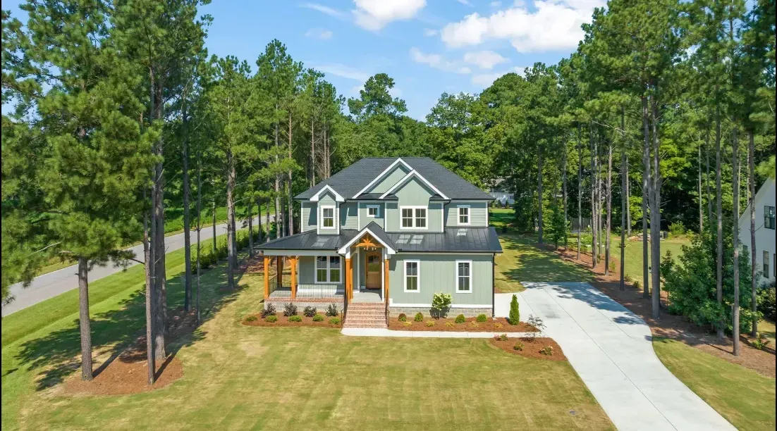 An aerial view of a sage green, two-story house with a wooden porch, surrounded by tall pine trees and a grassy yard.