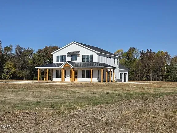 A two-story white house with a dark roof and a covered wooden porch sits on a cleared lot surrounded by autumn trees.