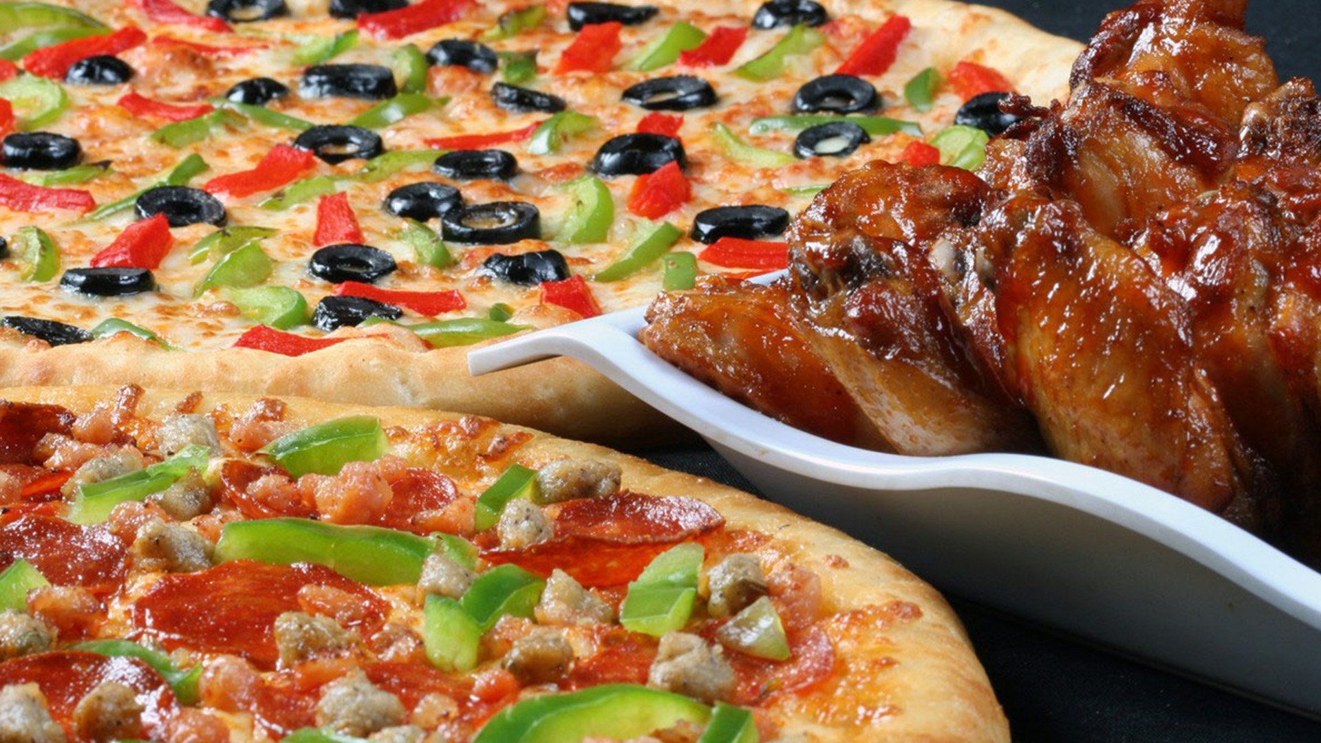 Two pizzas with various toppings and a side of glazed chicken wings.