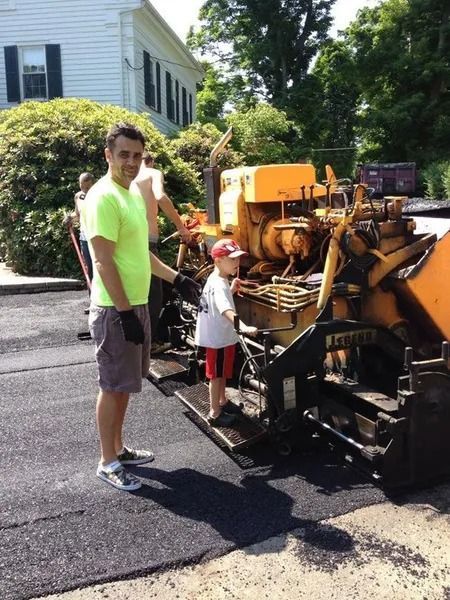Two people stand beside a yellow road machine on a residential street.