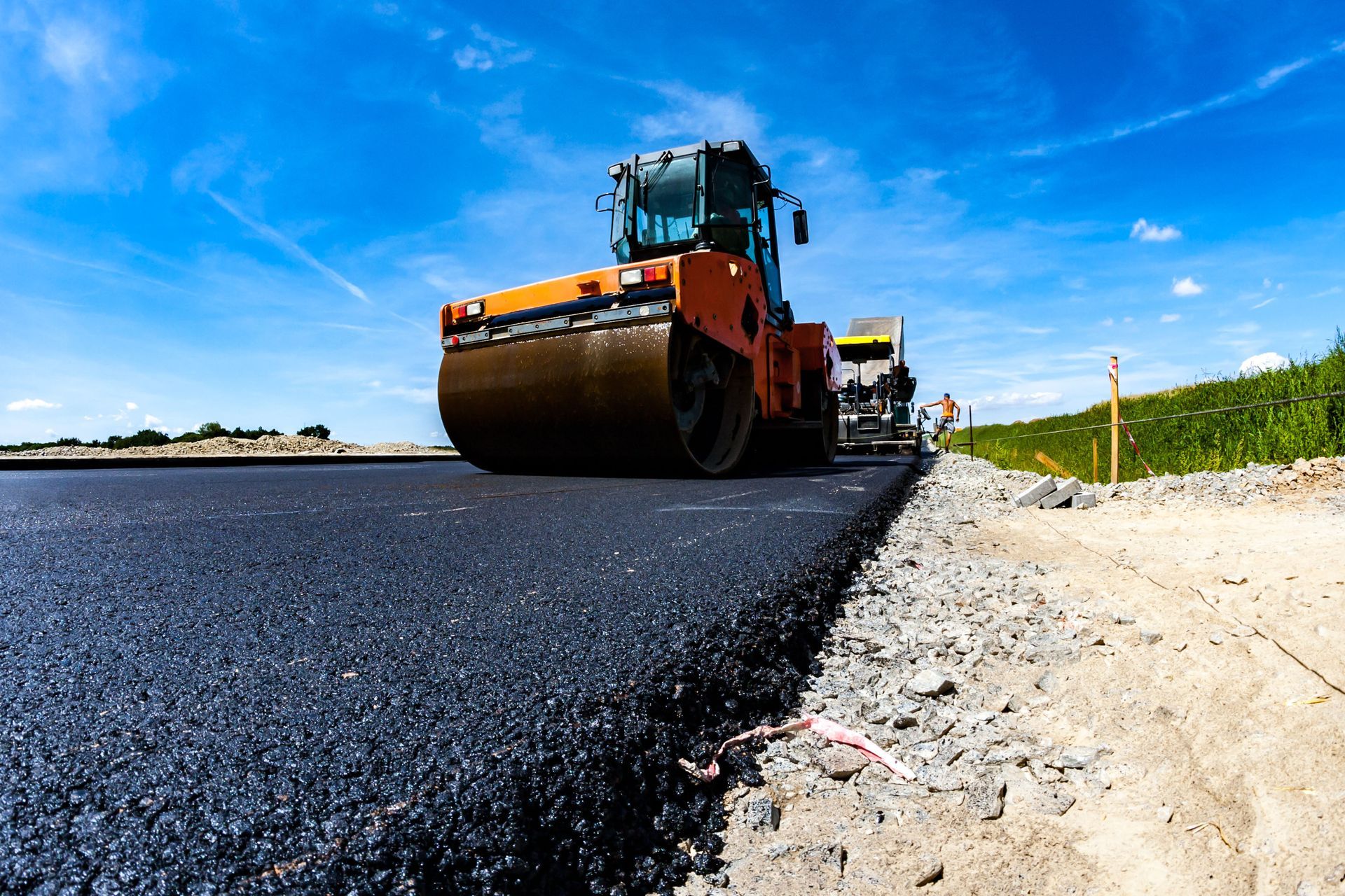 Road roller paving fresh black asphalt beside a sandy shoulder under a blue sky