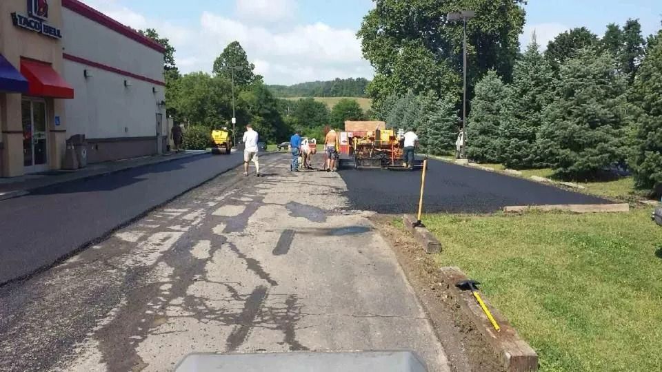 Road paving work near a building, with workers and equipment along a freshly resurfaced driveway.