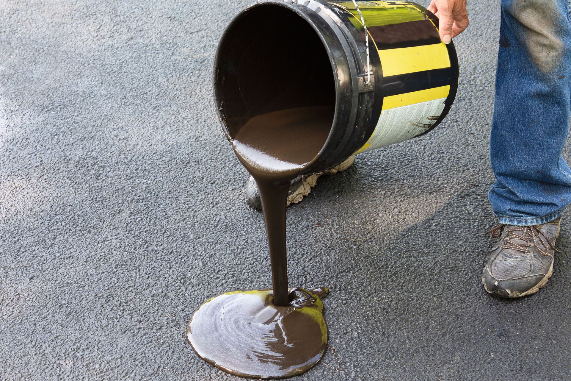 Worker pouring black asphalt from a bucket onto pavement, with yellow reflective stripes visible on the container.