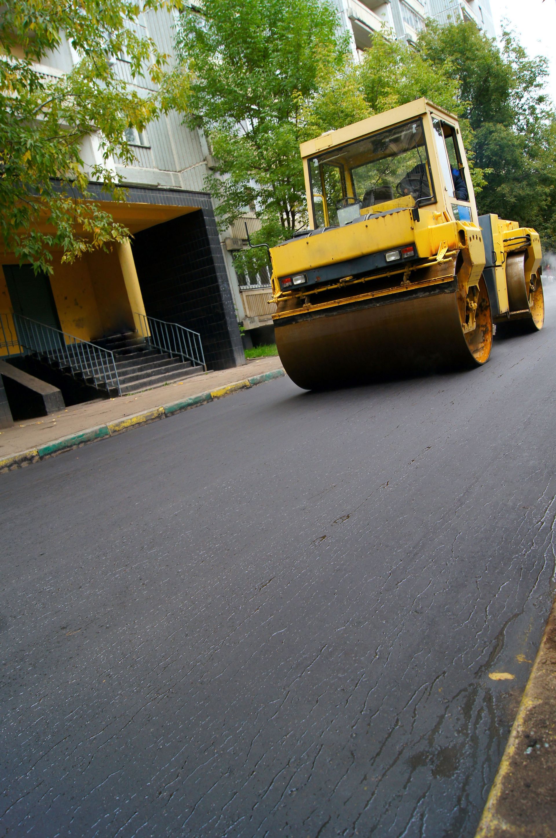 Yellow road roller paving a street beside a building and trees.