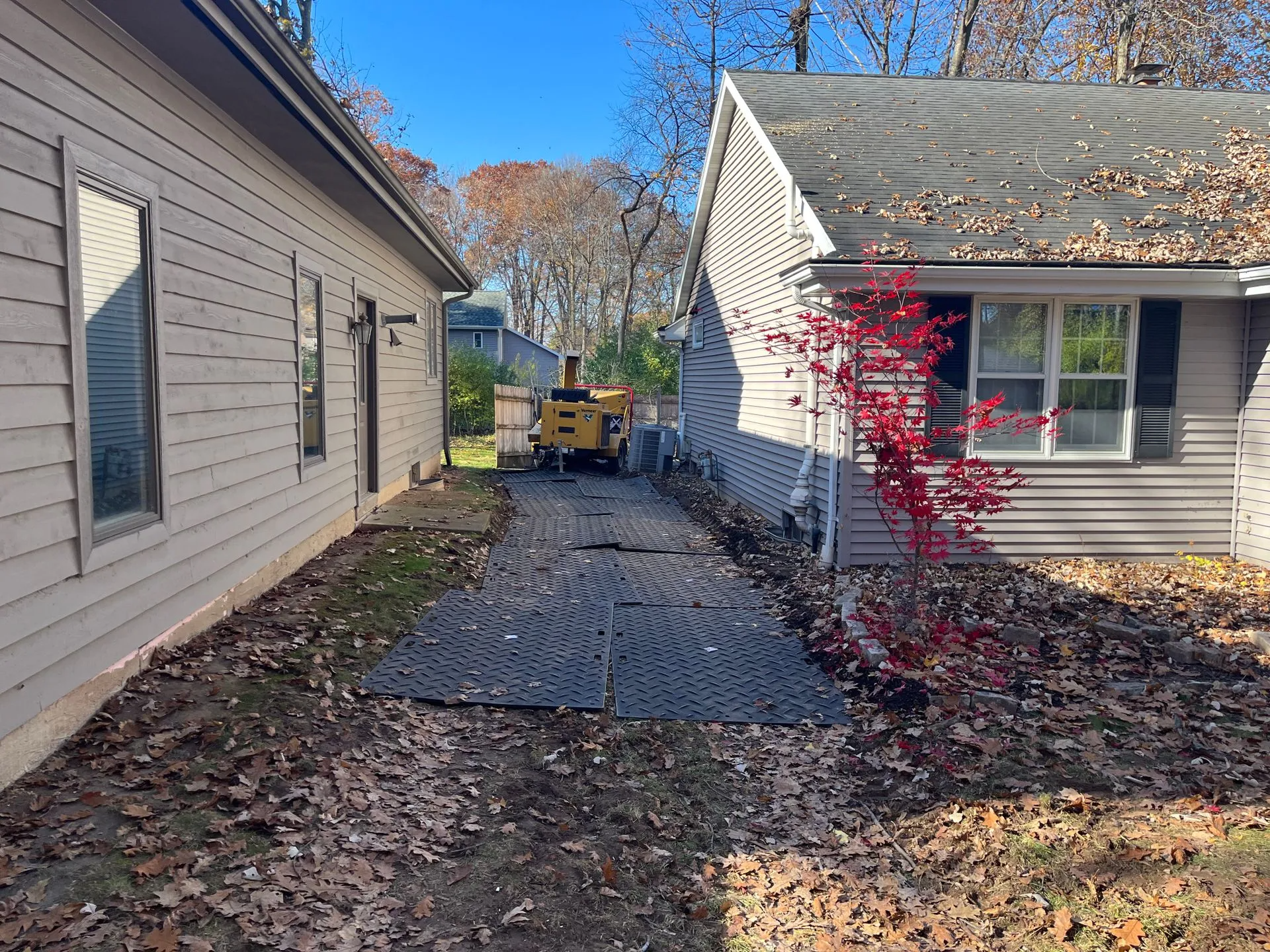 Backyard view: black mats on dirt path between two houses; a tree stump grinder is visible.