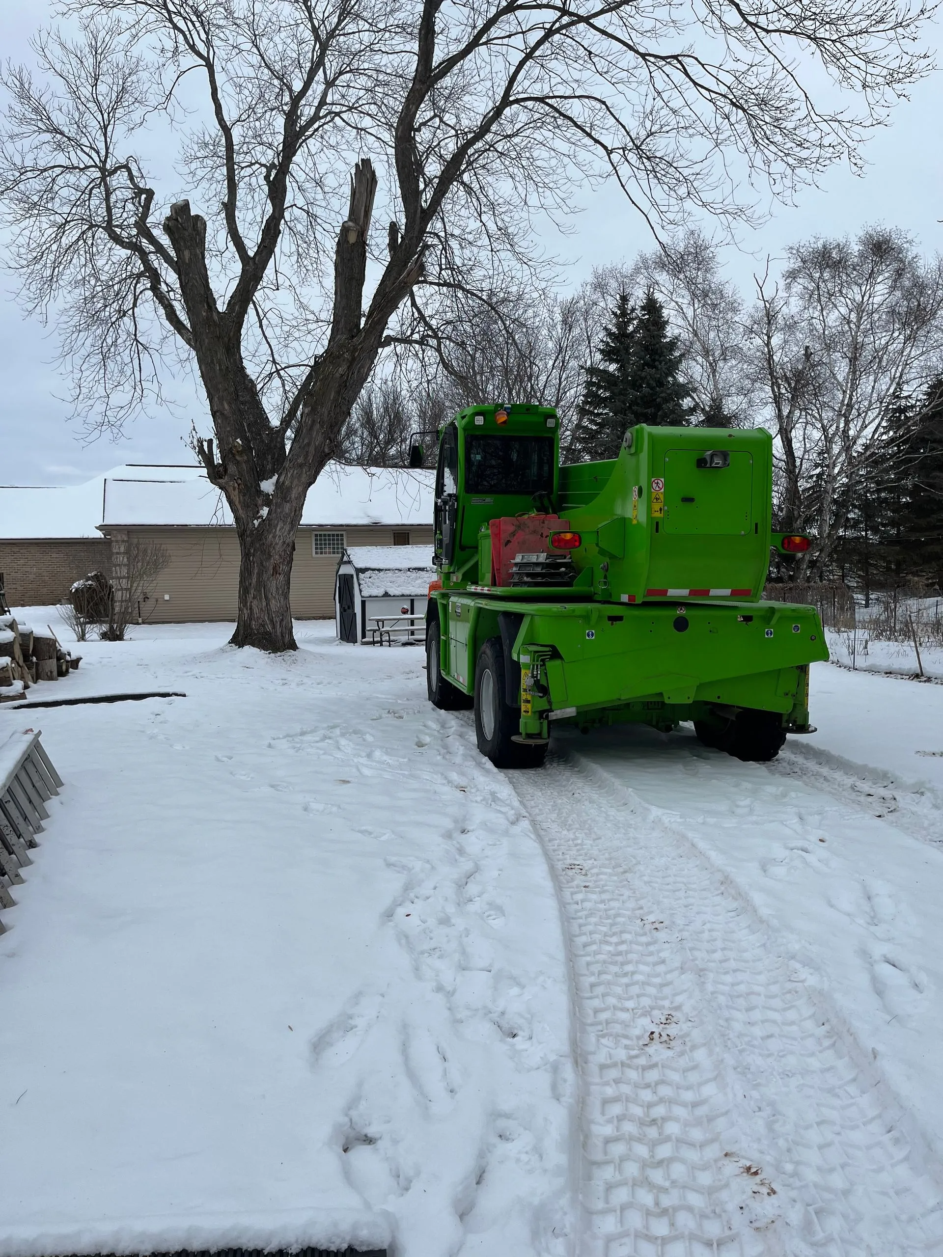 Green tree trimming truck in snow, near a house and partially pruned tree.