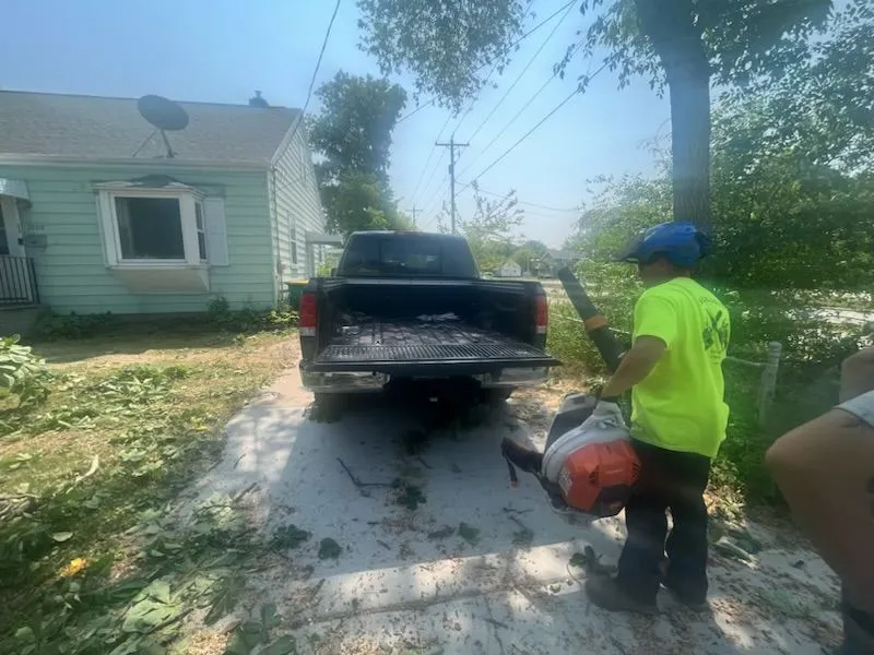 Person in neon shirt using a leaf blower, directing debris into a pickup truck on a driveway.