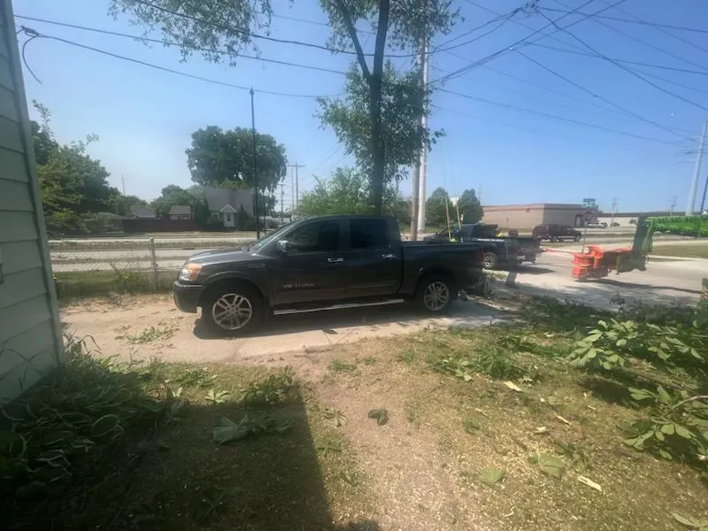 Dark pickup truck parked on a concrete slab next to overgrown grass, street in background.