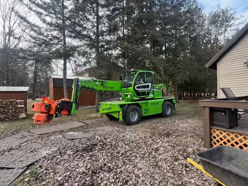 Green telehandler with a grapple attachment, next to a wood pile and house, lifting yard waste.