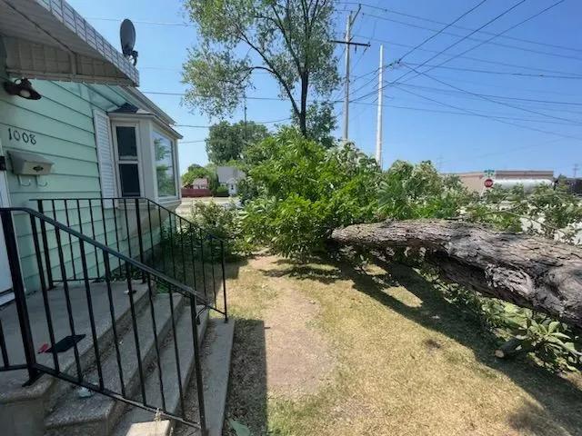 Fallen tree branch on lawn next to house with porch and steps. Green house and grass, blue sky.