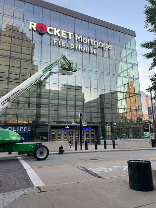 A construction lift parked in front of the glass-facade Rocket Mortgage FieldHouse building under a clear sky.
