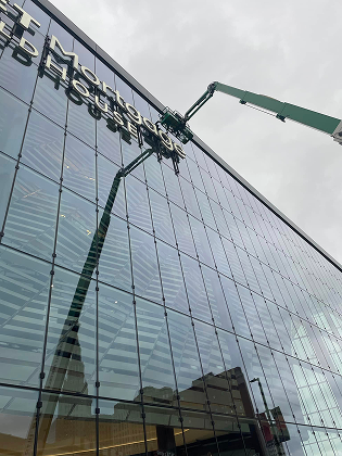 A crane arm reaches toward the top of a modern building with a glass facade, performing maintenance near signage.