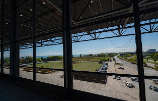A view from inside a building looking through large windows at a green lawn, parking lot, and distant coastline.