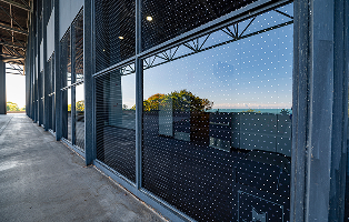 A row of tall glass panels with a white dot pattern reflects a sunny outdoor scene against a dark, industrial hallway.