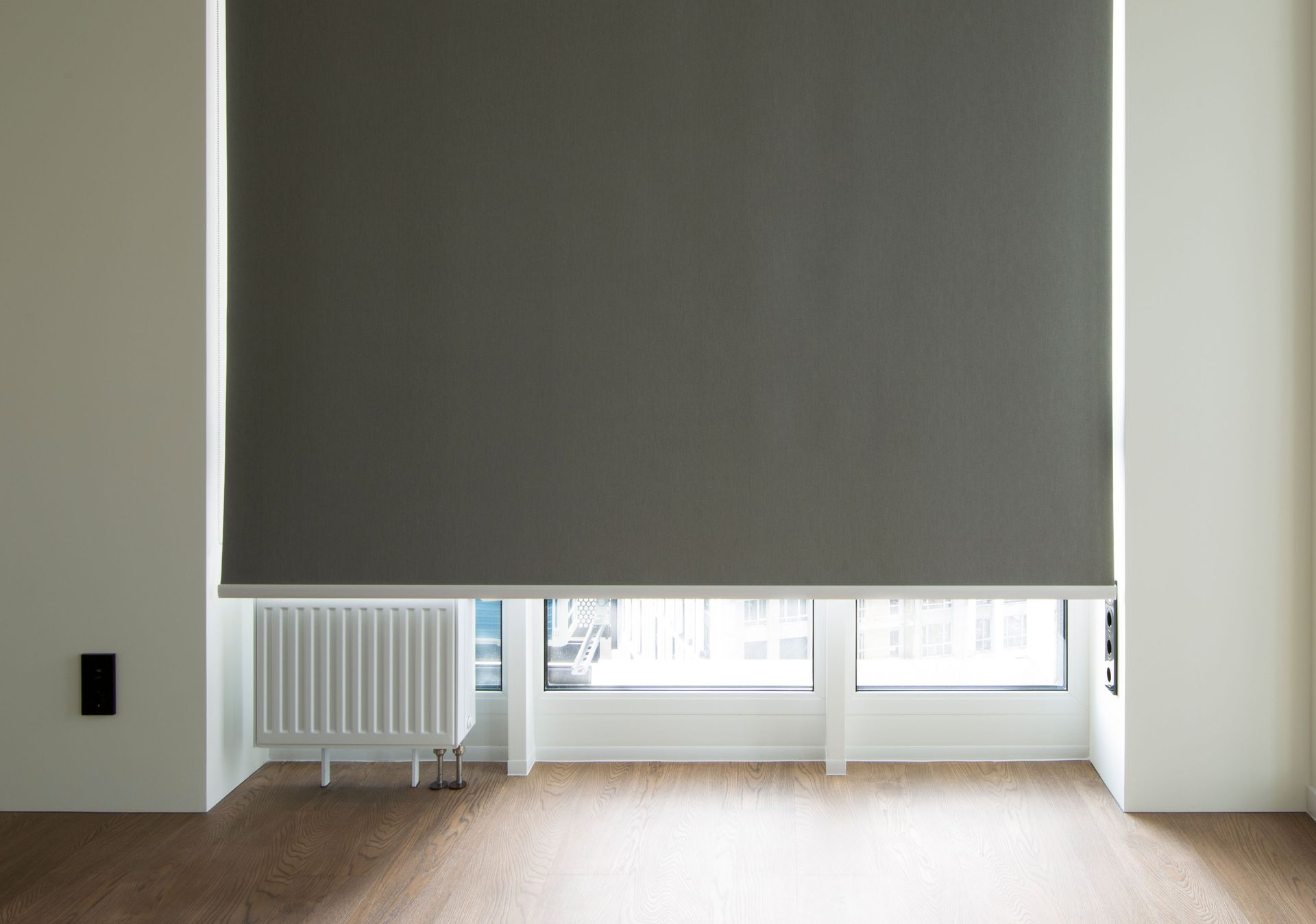 Gray roller shade covering a window in a bright room with a heater and wooden floor.