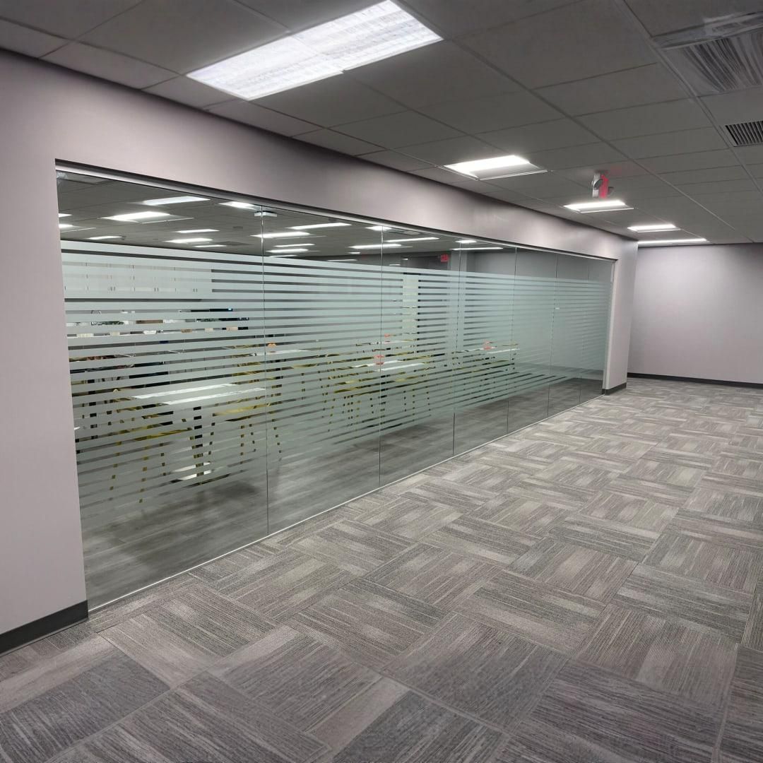 Empty office space with glass-walled section, gray carpet, and overhead lights.