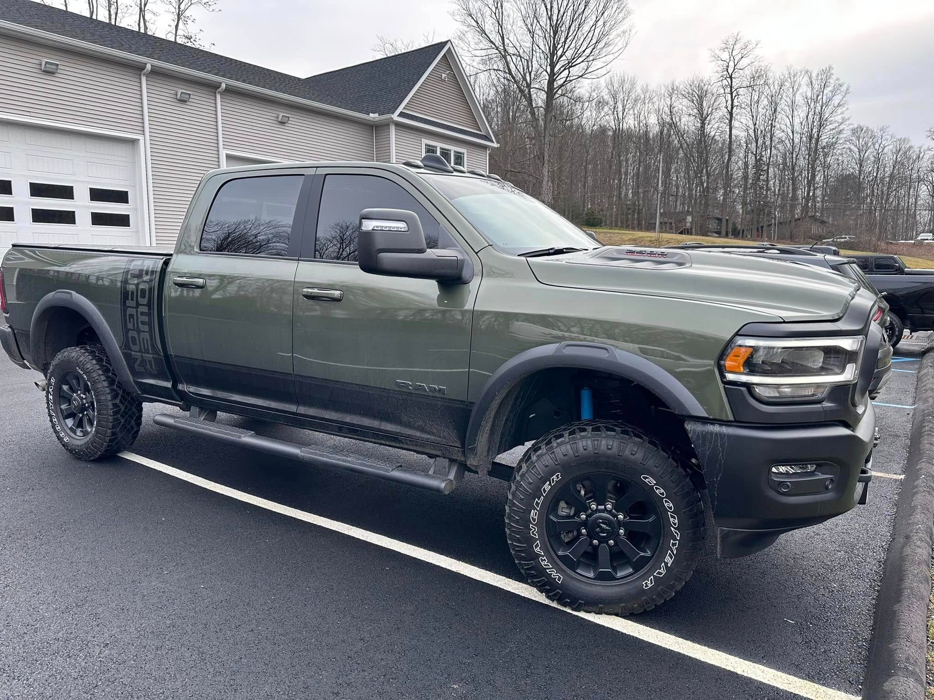 Green Ram Power Wagon truck parked on pavement. Black wheels, blue shocks, dark tint. Building and trees in the background.