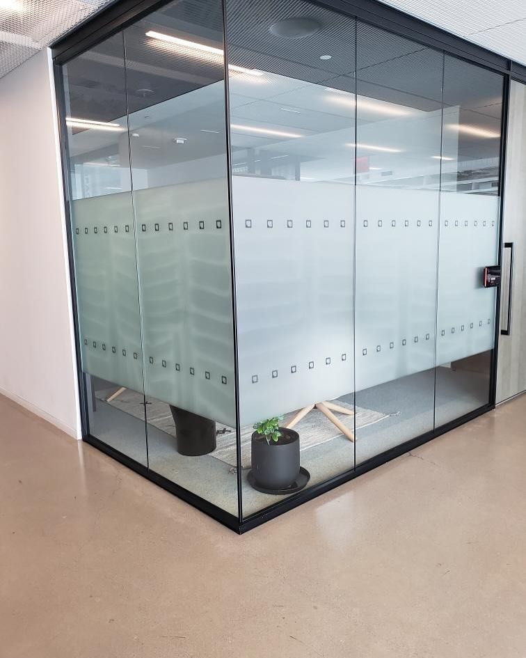 Glass-walled office corner with frosted privacy film, black door frame, and potted plants on the inside.
