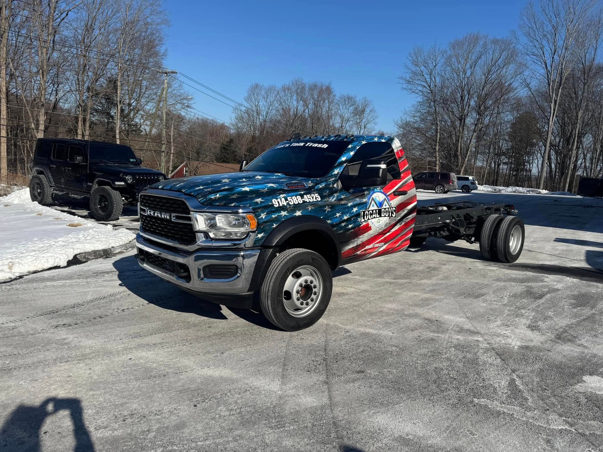 Ram truck with American flag graphics, parked on snow-covered ground. Jeep in the background. Sunny day.