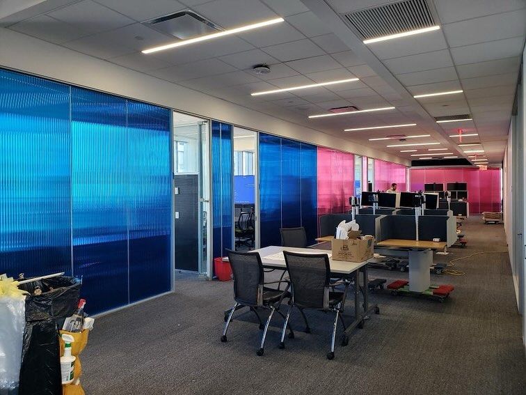 Office space with blue and pink translucent partitions. Desks, chairs, and construction materials are visible.