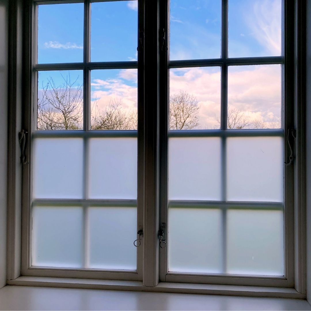 White-framed window with frosted glass. Outside is a blue sky with clouds and bare trees.
