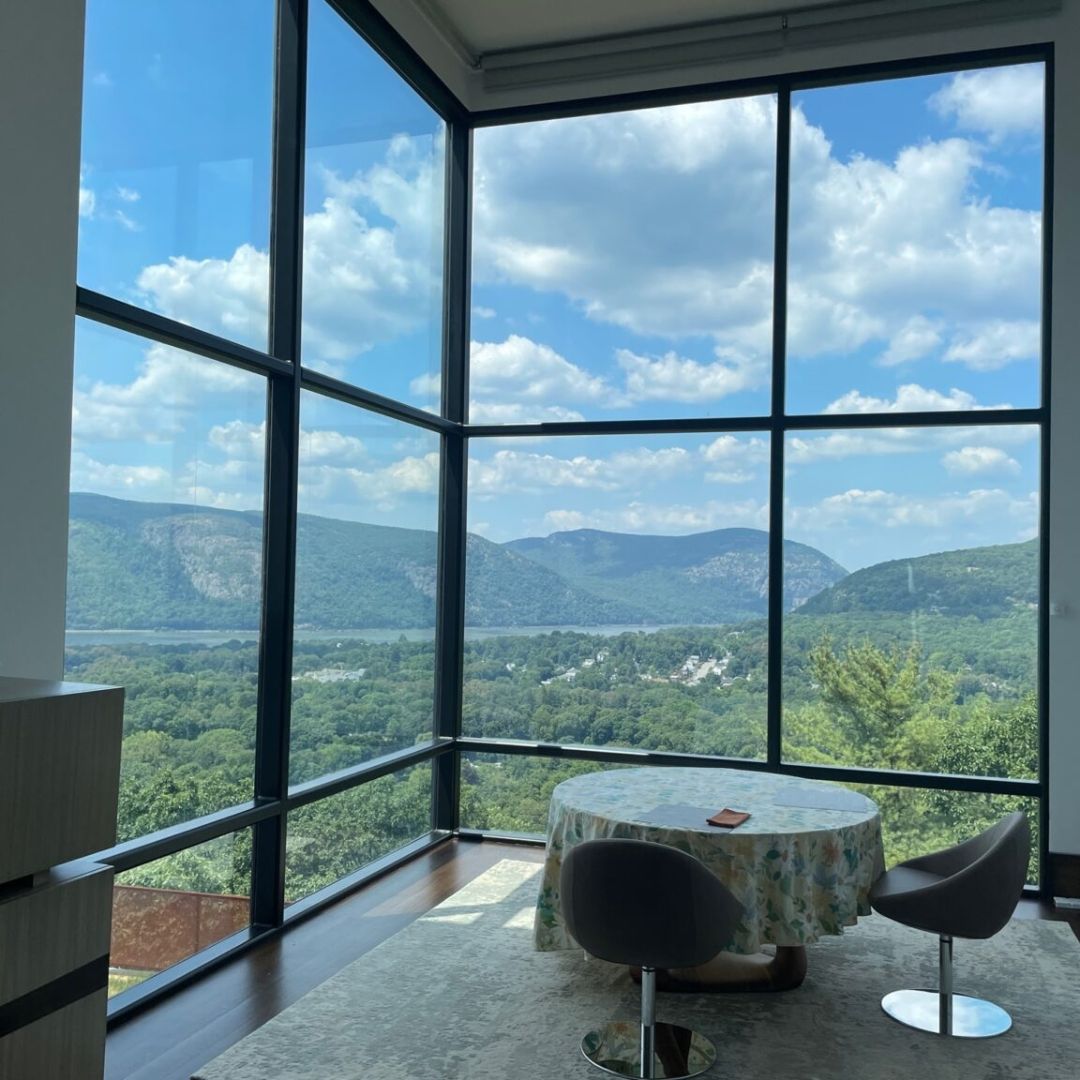 Corner room with large windows overlooking a green valley and mountains, round table and chairs.