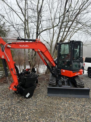 Orange Kubota mini excavator on gravel with a black attachment, parked outdoors among leafless trees.