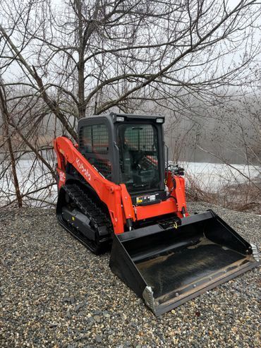 Orange skid steer with front bucket parked on gravel near leafless trees and a snowy field