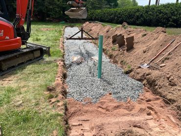 Excavator beside a gravel trench under construction, with a green pipe and dirt piles along a yard edge