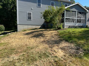 A sloped, mostly bare front yard with patchy grass beside a gray house and screened porch.