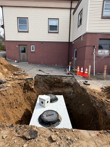 Construction site with a dug utility pit beside a building, exposed pipes, and orange safety cones.