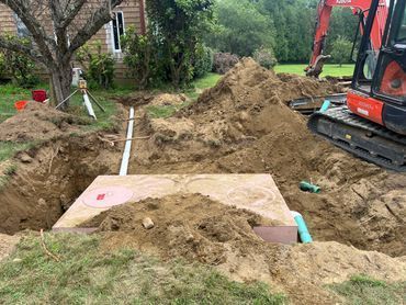 Backyard trench excavation with exposed pipes, dirt piles, and a small red excavator.