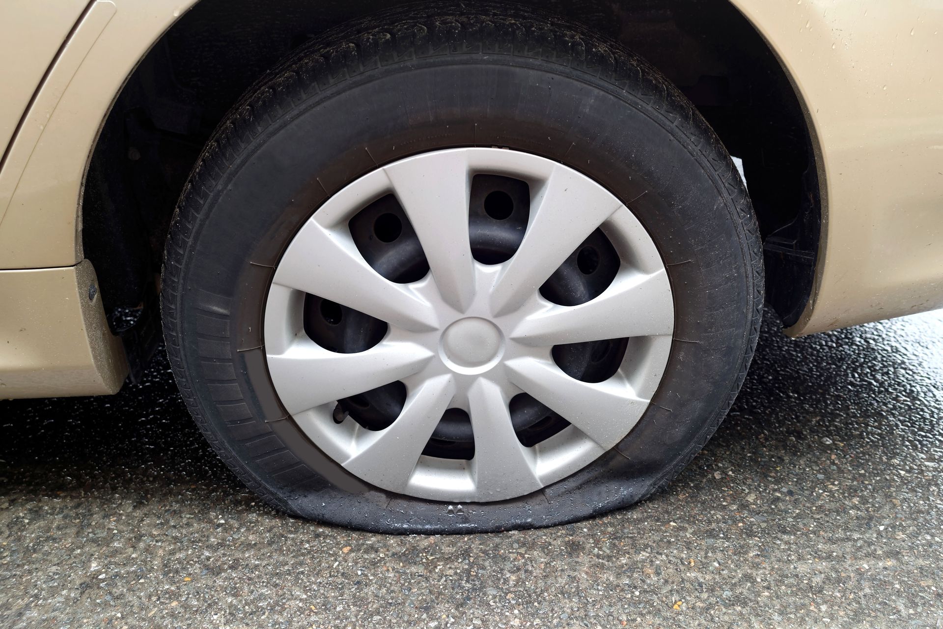 Flat car tire on a beige vehicle parked on wet pavement.