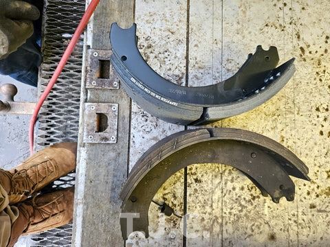 Two large curved black metal brake shoes beside a worker’s boots on a dirty shop floor