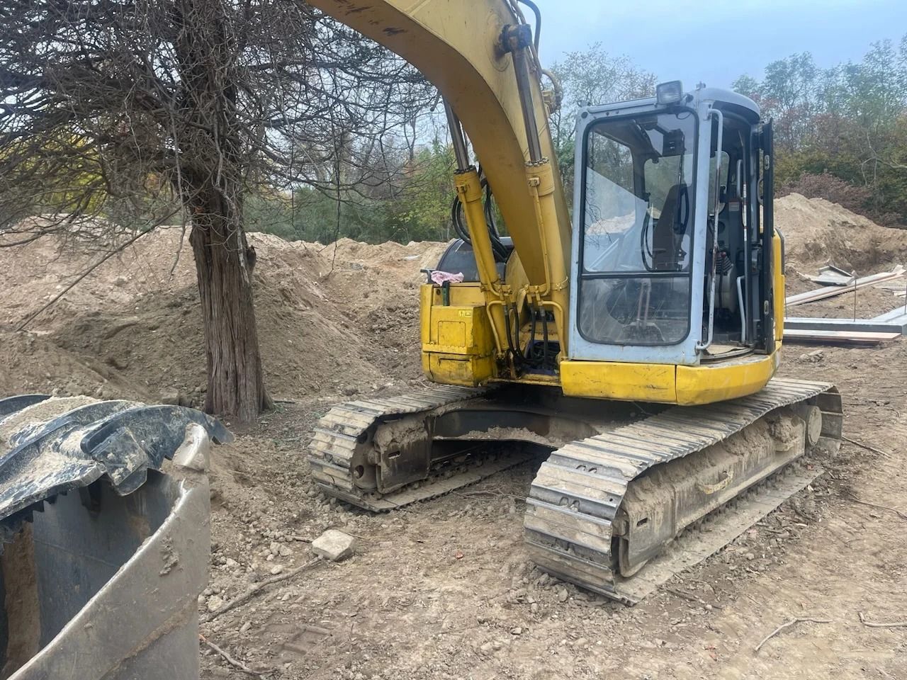 Yellow excavator on tracks beside a tree in a dirt construction site.