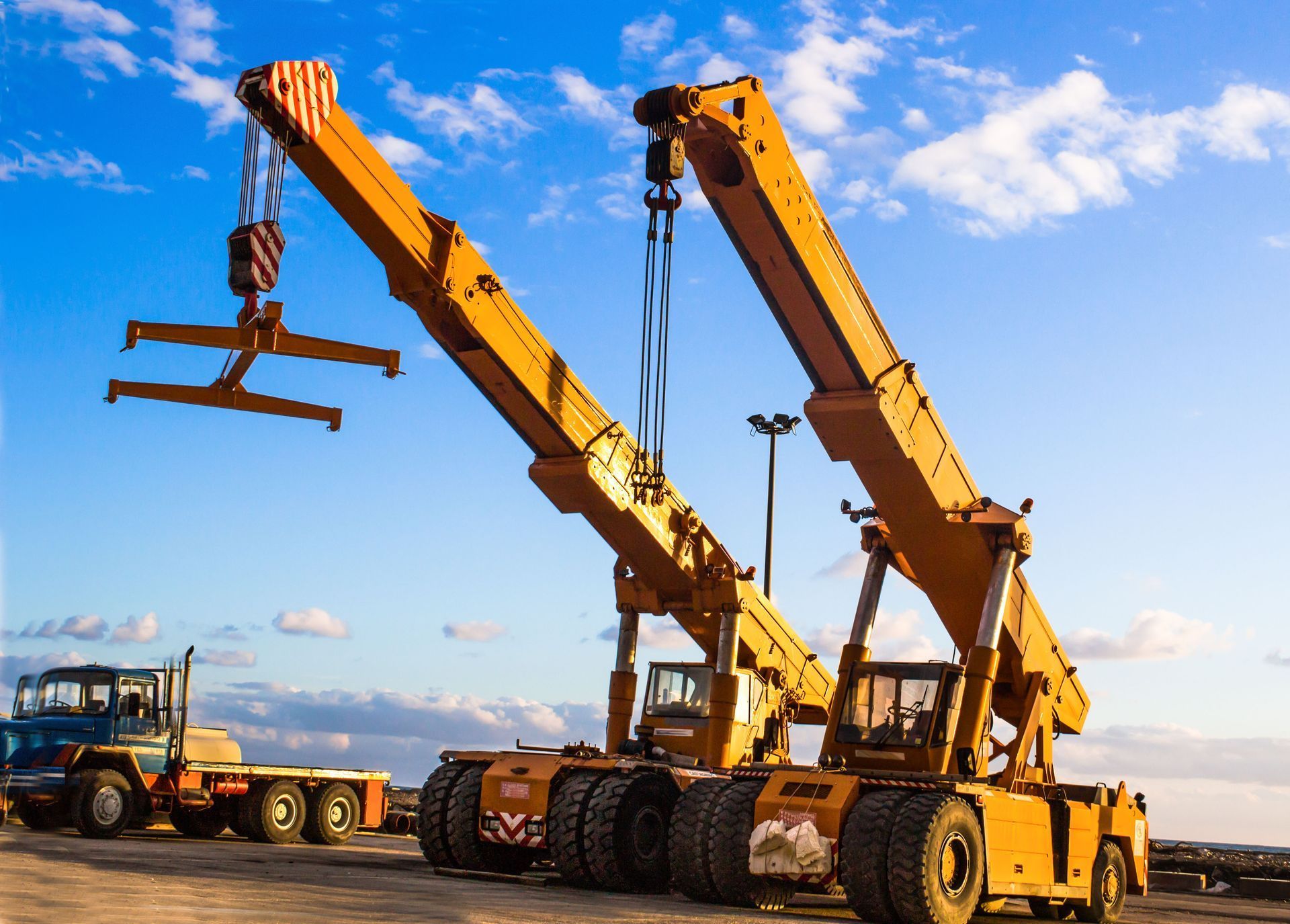 Two yellow mobile cranes sit side-by-side on a dirt lot under a blue sky, one lifting a spreader bar.
