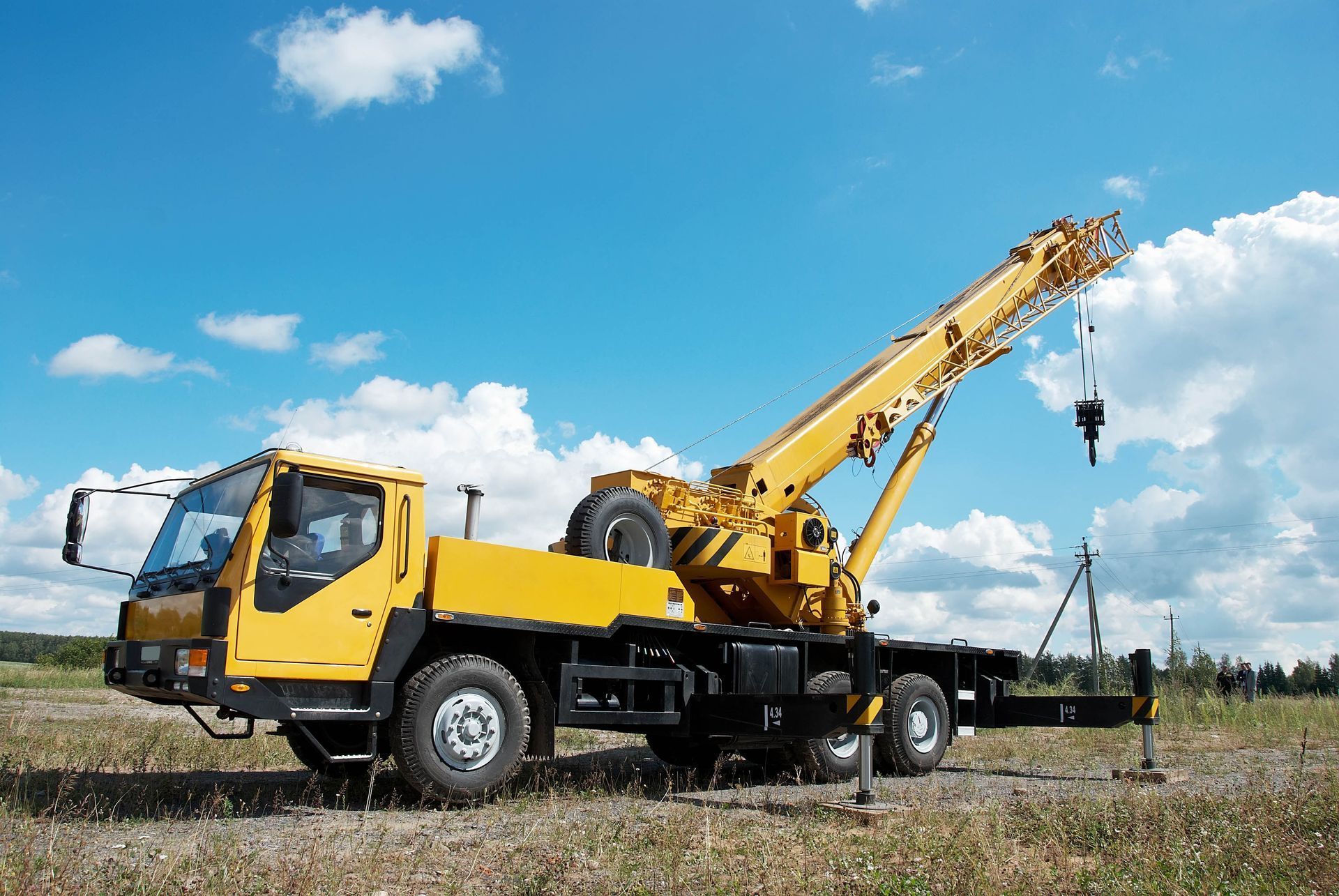 A yellow mobile construction crane parked on a gravel field under a bright blue sky with white clouds.