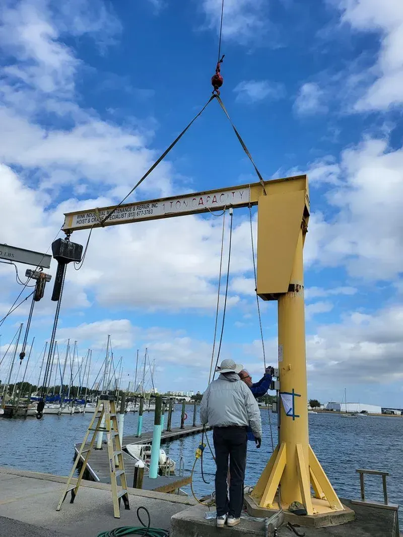 A person stands at a yellow jib crane on a dock by a marina, with the crane arm being lifted by an overhead hoist.
