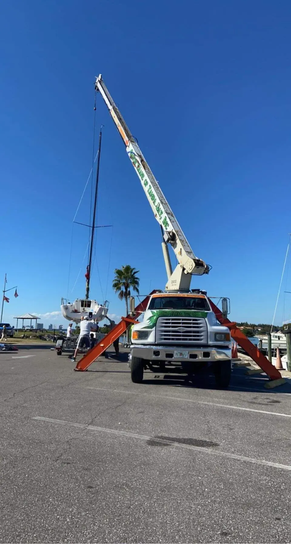 A large crane with extended outriggers is positioned in a parking lot, using its boom to lift a sailboat mast.
