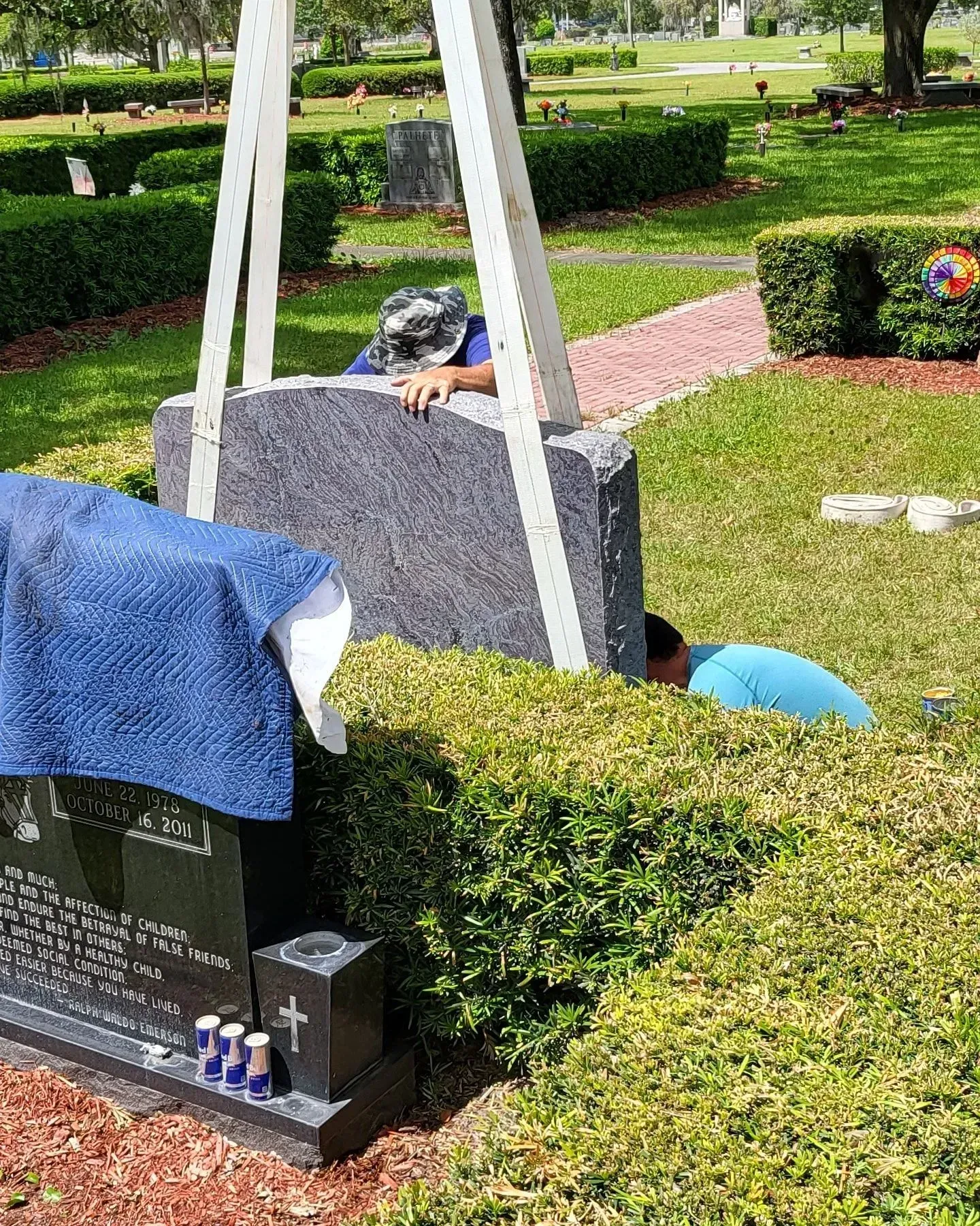 Two people work on installing or repairing a grey granite headstone in a cemetery on a sunny day.
