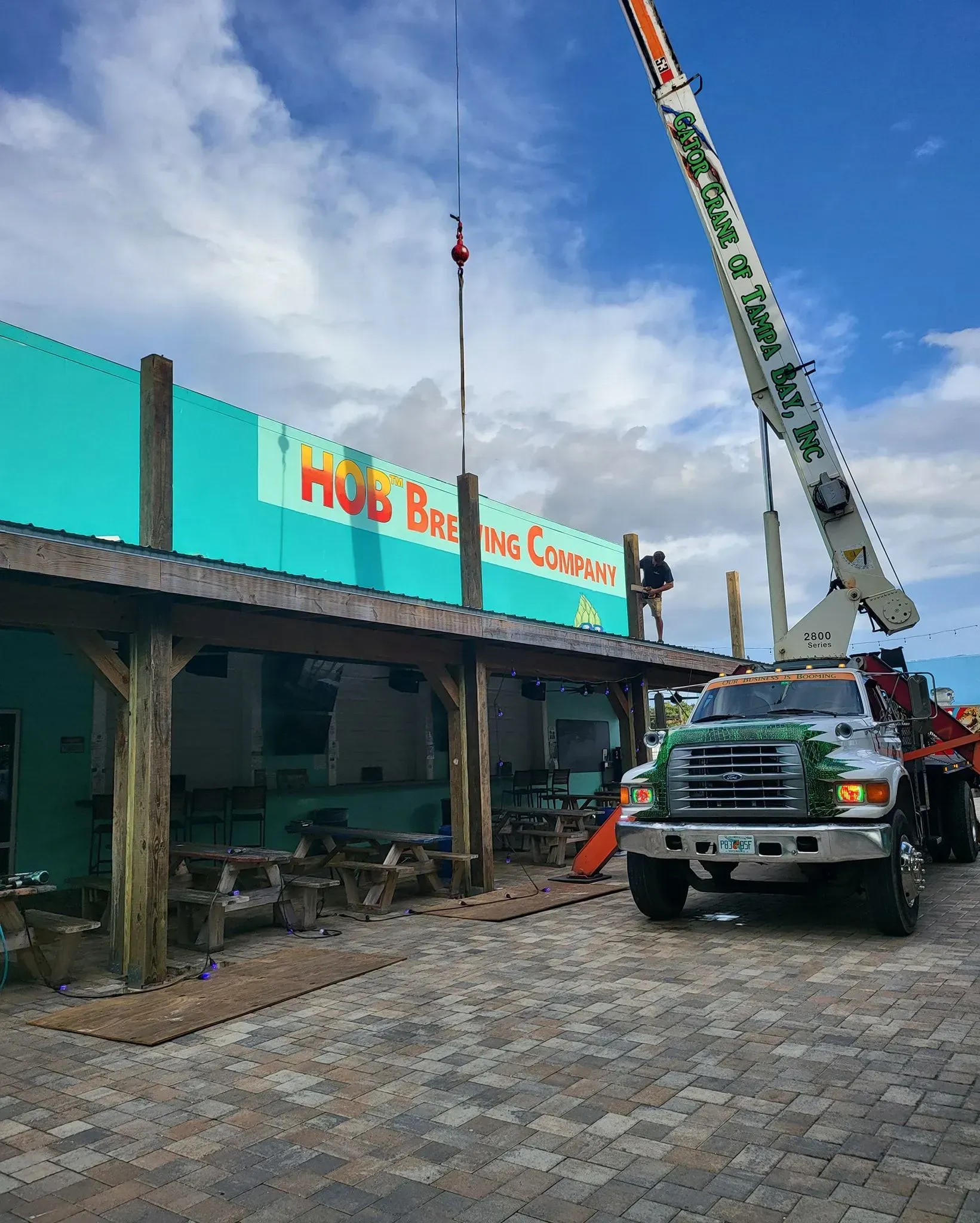 A crane truck parked next to a turquoise restaurant building with a wooden patio area under a partly cloudy blue sky.