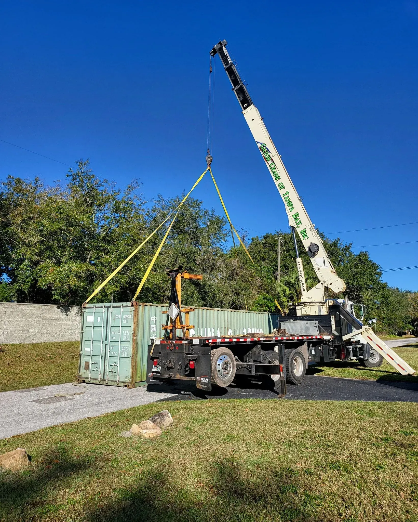 A crane lifts a green shipping container onto the bed of a truck parked on a grass field under a clear blue sky.