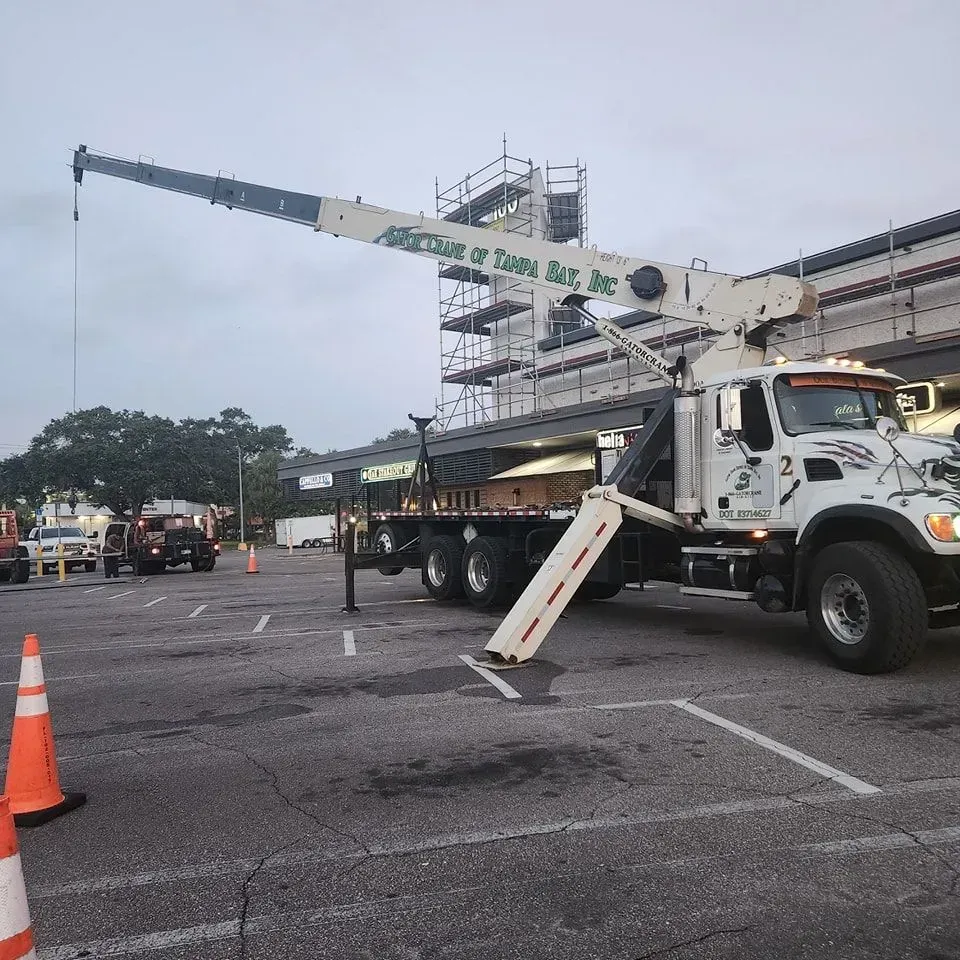 A white crane truck parked in a parking lot, with its extended boom pointed toward building scaffolding.