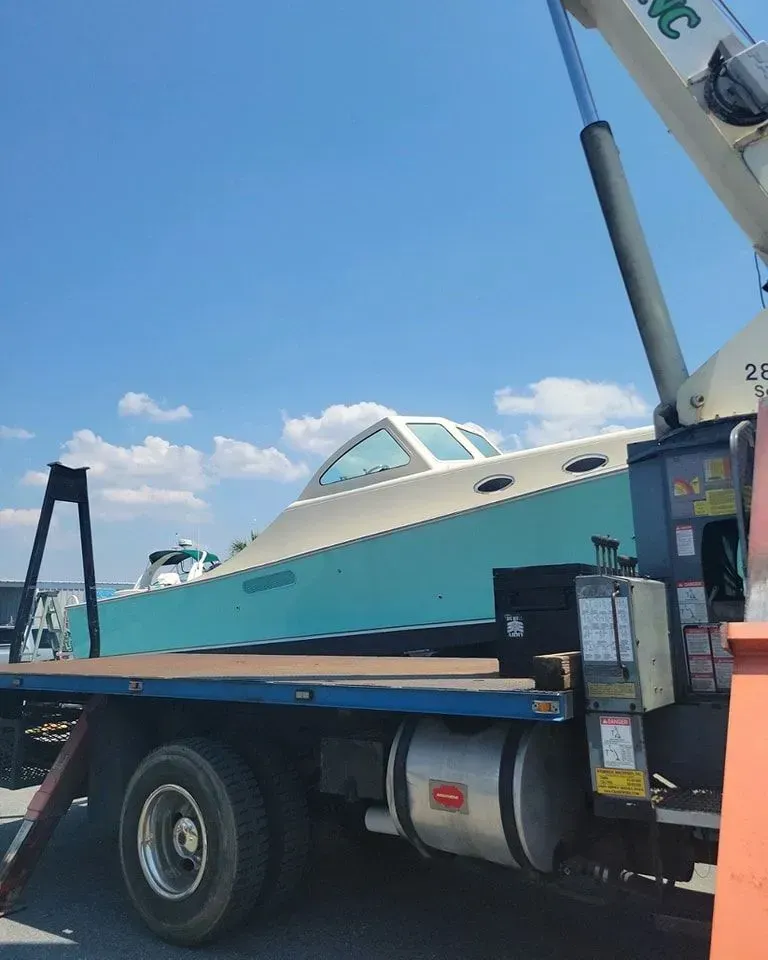 A teal and white cabin cruiser boat being loaded onto the flatbed of a crane truck under a blue sky.
