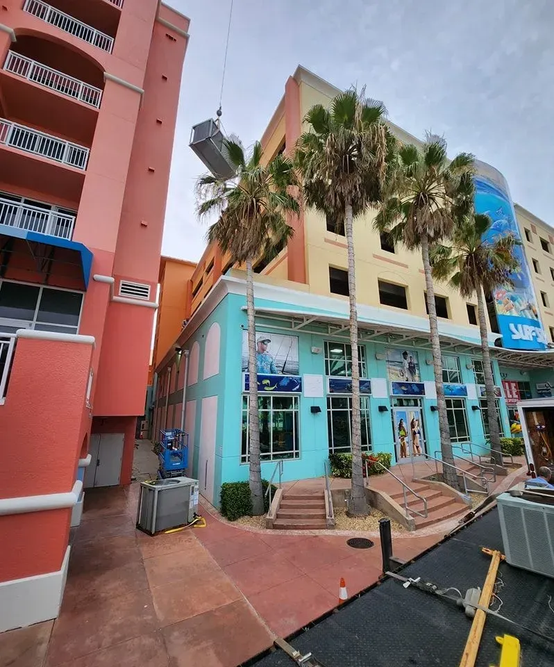 A crane hoists a large HVAC unit above a colorful commercial building complex with palm trees and a paved courtyard.