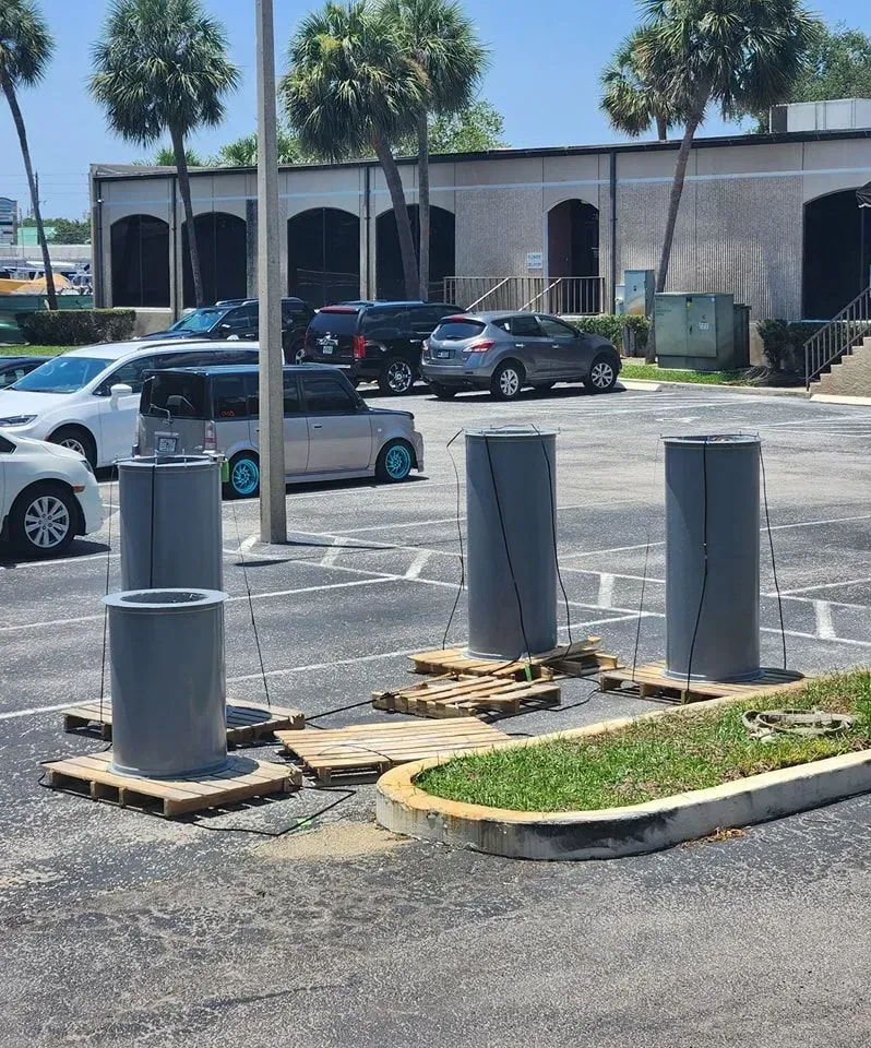 Four tall grey cylindrical pipes on wooden pallets sit in a parking lot with cars and a building in the background.