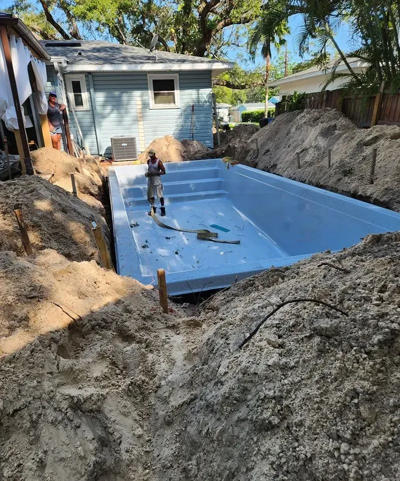 A worker installs a light blue, rectangular fiberglass swimming pool shell in a dirt excavation beside a residential house.